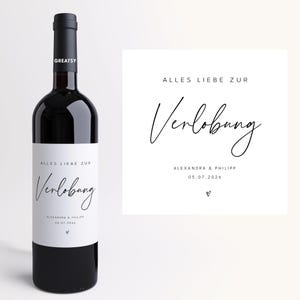 Könnte beinhalten: Eine dunkelrote Weinflasche mit einem weißen Etikett mit dem Text "ALLES LIEBE ZUR Verlobung" und den Namen "ALEXANDRA & PHILIPP" mit dem Datum "05.07.2026". Eine passende Karte befindet sich neben der Flasche.
