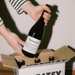 May include: A hand holding a bottle of red wine labelled "Patenonkel" with a white label. The bottle is being placed into a cardboard box with the word "Greatsy" printed on the side.