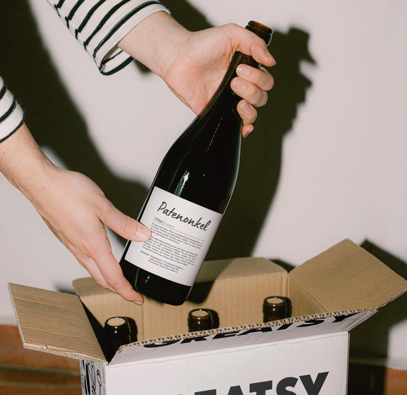 May include: A hand holding a bottle of red wine labelled "Patenonkel" with a white label. The bottle is being placed into a cardboard box with the word "Greatsy" printed on the side.