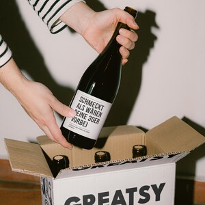 May include: A dark glass bottle with a white label is being placed into a cardboard box. The label reads "SCHMECKT ALS W&Auml;REN DEINE 30ER VORBEI". The box is labeled "GREATSY" in large black letters. Several other bottles are visible in the box.