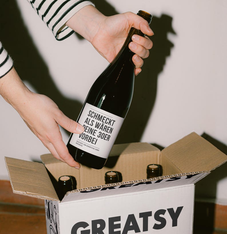 May include: A dark glass bottle with a white label is being placed into a cardboard box. The label reads "SCHMECKT ALS W&Auml;REN DEINE 30ER VORBEI". The box is labeled "GREATSY" in large black letters. Several other bottles are visible in the box.