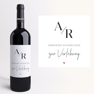Könnte beinhalten: Eine Flasche dunkelroten Weins mit einem weißen Etikett. Das Etikett zeigt die Buchstaben "A/R" und den deutschen Text "HERZLICHEN GLÜCKWUNSCH zur Verlobung" mit einem kleinen roten Herz. Die Flasche hat einen schwarzen Verschluss und die Marke "GREATSY".