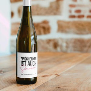 May include: A clear wine bottle with a white label featuring black and pink text that reads "EINSCHENKEN IST AUCH Schenken". The bottle is on a wooden surface. The text "HAPPY BIRTHDAY MONSCHT CHARLOTTE" is at the bottom of the label.