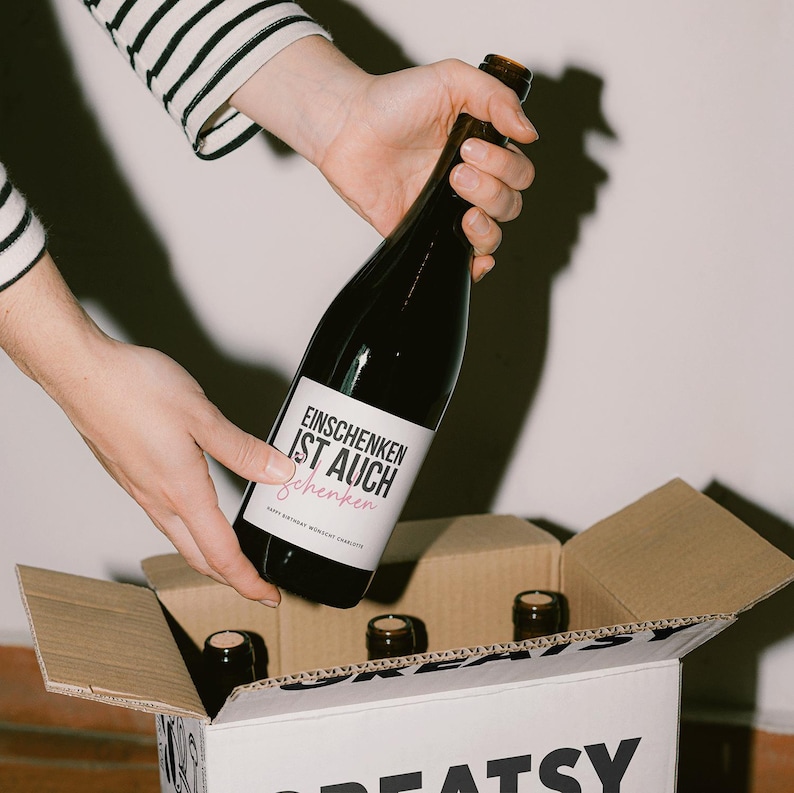 May include: A dark glass bottle with a white label featuring the text "EINSCHENKEN IST AUCH schenken" is held over a cardboard box containing other bottles. The label also includes the text "Happy Birthday Monscat Charlotte".