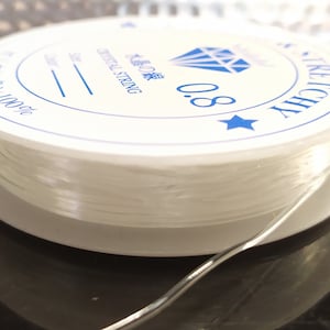 Può includere: Un rocchetto bianco di filo elastico trasparente con un'etichetta blu che dice "Jewelry String 0.8".