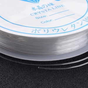 Peut inclure: Une bobine de fil de perles élastique transparent. L'étiquette présente du texte japonais et les mots "CRYSTA LINE" en bleu. Le cordon est enroulé serré sur une bobine blanche, avec une petite section de cordon qui s'étend.