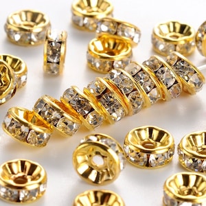 May include: A close-up of gold-toned metal bead caps with clear crystal rhinestones. The bead caps are arranged in a cluster on a white surface.