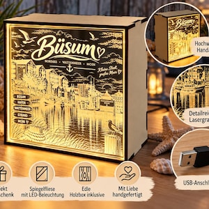 Könnte beinhalten: Eine rechteckige Holz-Lichtbox mit einer lasergravierten Szene der Stadt Büsum, einschließlich Hafen, Booten und Möwen. Die Frontplatte ist mit warmem LED-Licht beleuchtet und zeigt den Text Büsum, Nordsee, Wattenmeer, Moin.