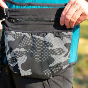 Puede incluir: Una riñonera de camuflaje negro y gris con un bolsillo con cremallera y una correa negra. La riñonera se lleva alrededor de la cintura y tiene un pequeño logotipo negro y verde que dice "GOX".