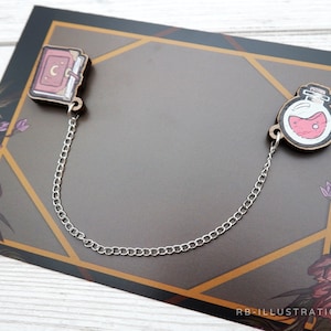 May include: A silver chain connects two wooden charms, one depicting a closed book with a crescent moon and the other a potion bottle with pink liquid. The charms are on a brown background with floral designs.