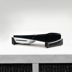May include: A white and black gaming console resting horizontally on a black plastic stand with a vented design. The console features a sleek, curved exterior with a glossy black center panel and white side plates, accompanied by two controllers.