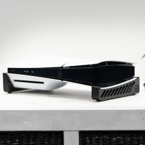 Puede incluir: Una consola PlayStation 5 blanca con un soporte negro. La consola está sobre una superficie blanca y está ligeramente inclinada hacia arriba. El soporte tiene un diseño negro y gris.