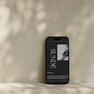 May include: A black smartphone displays a dark screen with the word "SUNDË" in white, along with the text "THE REBEL" and a grayscale image. The phone is set against a neutral background with leaf shadows.
