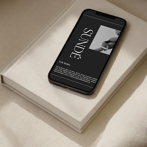 May include: A black smartphone with the word "SUNDE" in white, a monochrome image, and text, sits on a white book. The book is on a beige textured surface, creating a minimalist aesthetic.