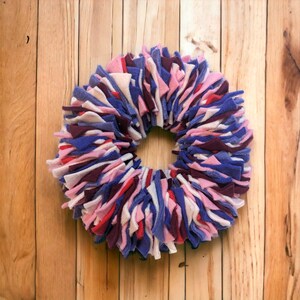 May include: A wreath made of strips of fabric in shades of pink, purple, white, and red. The wreath is made of felt and is a circular shape.