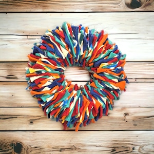 May include: A colorful, handmade wreath with a circular design. The wreath is made of strips of fabric in various colors, including blue, orange, red, and yellow. The wreath is displayed on a wooden background.