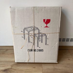 May include: A cardboard box tied with twine, featuring a red broken glass symbol and a line drawing of nesting tables. The text "ITEM NO.31885" is printed on the box. The box is on a wooden floor.