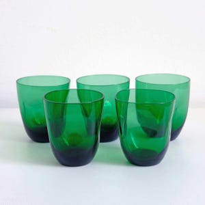 Può includere: Un set di cinque bicchieri in vetro verde smeraldo con una silhouette liscia e arrotondata. Ogni bicchiere presenta una tonalità verde traslucida e vibrante che si scurisce verso la base, con un design semplice senza stelo e bordo ampio.