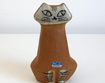 May include: A brown ceramic cat figurine with a light gray face and black facial details. The cat has a long, slender body and is seated. The figurine has a small blue and white sticker that reads "Design Lisa Larson".