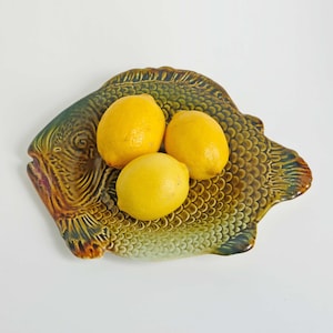 Könnte beinhalten: Eine Keramik-Servierplatte in Fischform mit einer strukturierten Schuppenoptik in Grün-, Braun- und Gelbtönen. Die Platte hat eine glänzende Oberfläche und trägt drei leuchtend gelbe Zitronen auf ihrer Fläche, was ihr funktionales Design unterstreicht.