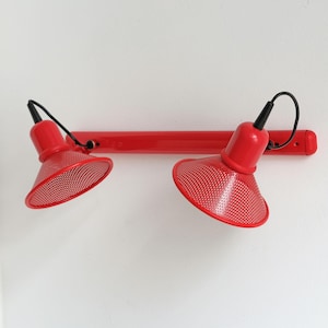 May include: A red wall light with two adjustable lamps. Each lamp has a perforated shade and a black cable. The fixture is mounted on a white wall.