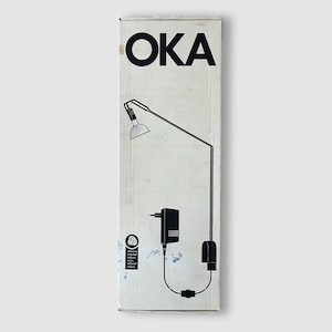 Può includere: Illustrazione in bianco e nero di una lampada da scrivania su una scatola di cartone. La scatola ha la parola "OKA" in grandi lettere nere in alto. La lampada ha un design minimalista con un braccio lungo e sottile e un piccolo paralume rotondo.