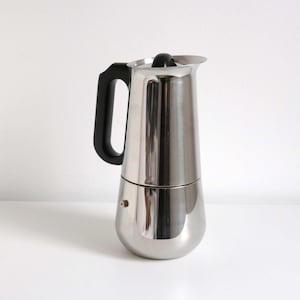 Pode incluir: Uma cafeteira espresso de aço inoxidável polido com corpo cilíndrico cônico, alça preta resistente ao calor e um botão preto correspondente na tampa articulada. A base é arredondada e possui uma pequena válvula de pressão metálica.