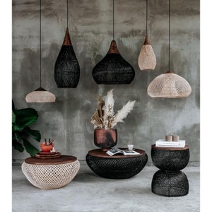 K&ouml;nnte beinhalten: Ein Wohnzimmer-Interieur mit einer Auswahl an geflochtenen Pendelleuchten und geflochtenen Couchtischen. Die Leuchten sind schwarz und naturfarben, die Tische sind schwarz und naturfarben. Auf einem der Tische steht eine Vase mit Trockenblumen.