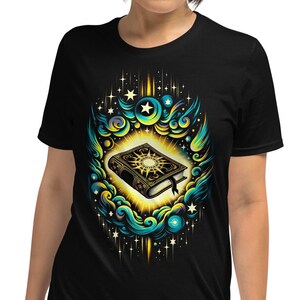 Puede incluir: Una camiseta negra con un gráfico de un libro rodeado de nubes, estrellas y un sol brillante. El libro está abierto y tiene un diseño dorado.