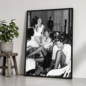 May include: A black and white framed wall art print depicting a backstage scene with several individuals preparing for an event. The composition features people in casual clothing and hair rollers, captured in a candid moment within a room.