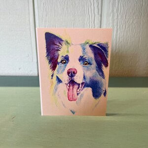 May include: A watercolor painting of a border collie dog with blue, purple, and green hues. The dog is smiling with its tongue sticking out.