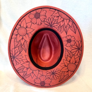 May include: A wide-brimmed hat in a vibrant coral-red hue, featuring a black interior band and intricate black line-art illustrations of various sunflowers and daisies covering the entire upper surface of the brim and crown.