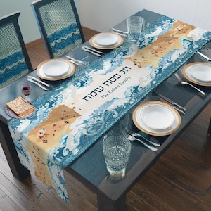 May include: A blue and white table runner with a wave pattern and a gold inscription that reads "Happy Passover The Cohen Family". The runner is laid out on a wooden table with place settings.