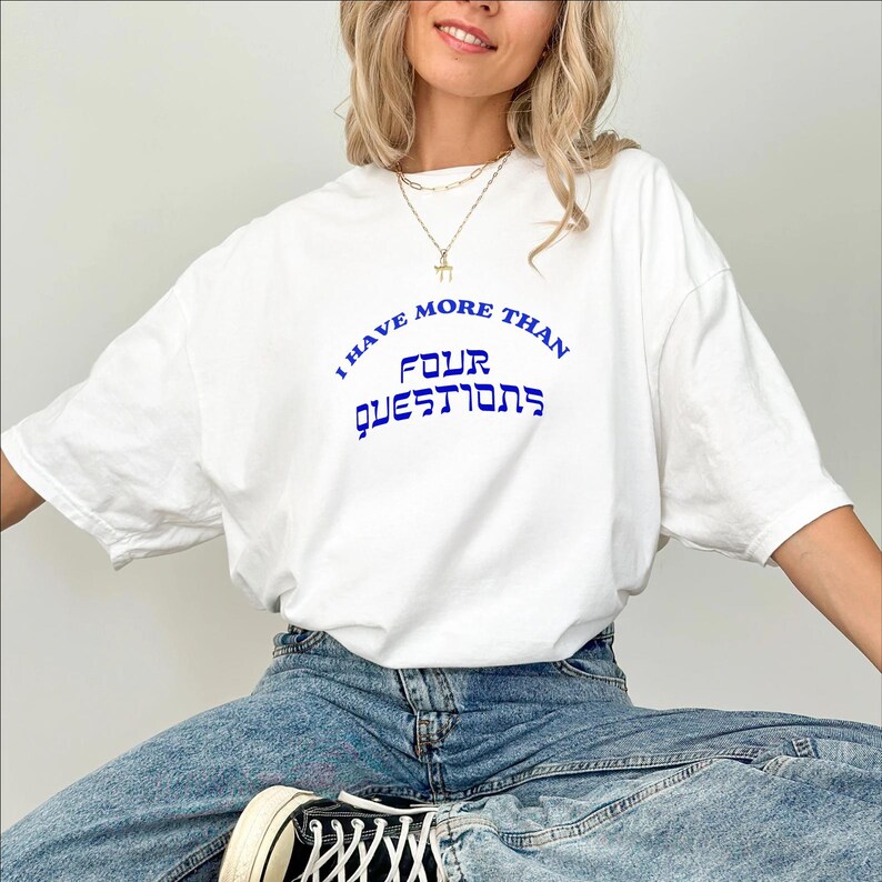 May include: White t-shirt with the blue text "I HAVE MORE THAN FOUR QUESTIONS". The shirt is a casual style, with a relaxed fit. The text is in a stylized font. The model is wearing blue jeans and black sneakers.