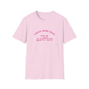 May include: A light pink t-shirt with the text "I HAVE MORE THAN FOUR QUESTIONS" in a darker pink, arched above and below the words. The shirt has short sleeves and a crew neck.