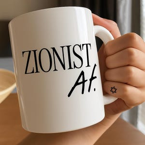 Puede incluir: Taza de cerámica blanca con el texto "ZIONIST Af." en negro. La taza está sujeta por una mano con un pequeño tatuaje de la Estrella de David. La taza tiene un asa curvada.