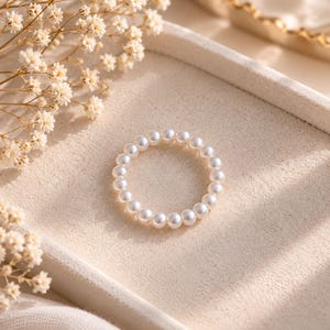 May include: A circular ring composed of small, uniform white spherical pearls linked together in a continuous band. The pearls have a smooth, lustrous finish and are arranged in a simple, unbroken loop, creating a delicate and refined jewelry piece.