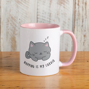 Puede incluir: Una taza de cerámica blanca con un borde y un asa de color rosa. La taza presenta un gato gris de dibujos animados durmiendo con el texto "Napping is my cardio" impreso en la parte delantera.