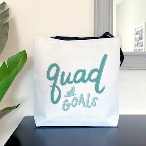 May include: White tote bag with black handles featuring the text "Quad GOALS" in teal script and a teal roller skate graphic. The bag is sitting on a dark surface.