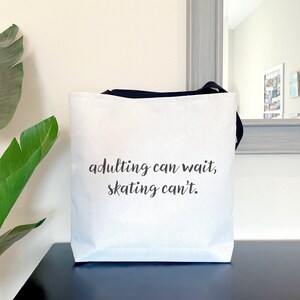 May include: White tote bag with black handles and the text "adulting can wait, skating can't." The bag is sitting on a dark surface with a large green plant to the left. A mirror is in the background.