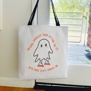 May include: White tote bag with black straps, featuring a cartoon ghost wearing ice skates. The text reads, "Nothing spookier than hitting the ice with your skate guards on." The design is in orange and black.