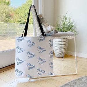 May include: White canvas tote bag with blue ice skate print. The bag has black handles and is sitting on a white chair.