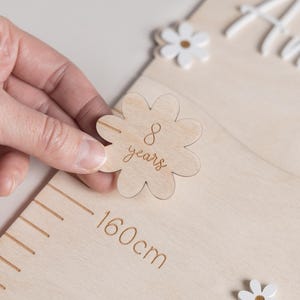 May include: A wooden height chart with a ruler marked up to 160cm. A hand holds a wooden flower-shaped marker that says "8 years". The name "Amelia" is written in white script. White daisy accents are scattered around the chart.