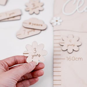 Op de afbeelding: Houten groeimeter met de naam "Clara" en madeliefjesaccenten. Inclusief houten bloemvormige markeringen voor de leeftijden 2, 3, 4, 7 en 8 jaar. De meter heeft een schaal tot 160 cm.