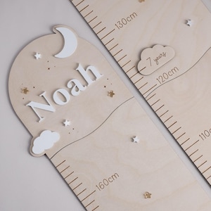Può includere: Grafico di crescita in legno chiaro, con luna, stelle e il nome "Noah" in bianco. Include una nuvola con "7 anni" e misurazioni in centimetri fino a 160cm. Decorativo e funzionale per monitorare l'altezza di un bambino.