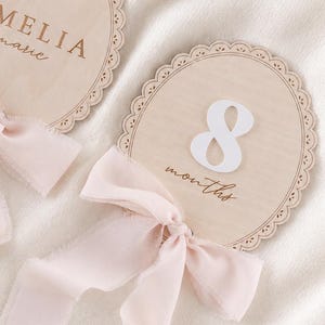 Könnte beinhalten: Zwei ovale Holzschilder mit gewelltem Rand und hellrosa Schleifen. Ein Schild trägt den Text AMELIA marie in Braun, das andere zeigt die Zahl 8 in Weiß mit dem Wort months in brauner Schreibschrift.