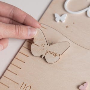 Op de afbeelding: Een houten groeimeter met een vlindervormige markering met de tekst "5 jaar". De meter heeft een liniaal met metingen in centimeters en een decoratieve vlinder en naam in het wit. De meter is gemaakt van lichtgekleurd hout.