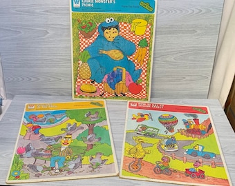 May include: Three vintage Sesame Street frame-tray puzzles. The first puzzle features Cookie Monster having a picnic. The second puzzle shows Big Bird sitting on a bench with pigeons. The third puzzle shows Big Bird, Elmo, and Cookie Monster on a Sesame Street bus.
