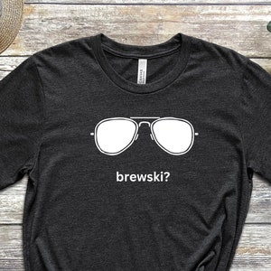 Puede incluir: Una camiseta de color negro jaspeado con un gr&aacute;fico blanco de gafas de sol y el texto "brewski?" impreso en la parte delantera.