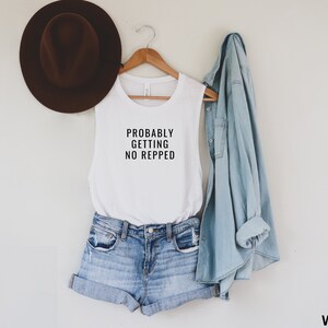 Op de afbeelding: Een wit tanktop zonder mouwen met de tekst "Probably Getting No Repped" erop gedrukt. Het tanktop wordt gemodelleerd door iemand die een spijkerbroek en een spijkerjack draagt.